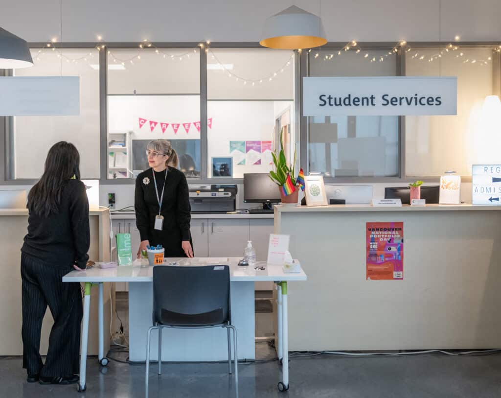 Two people talk at the Student Services front desk, which is decorated with small lights, posters, and plants inside a bright campus office.