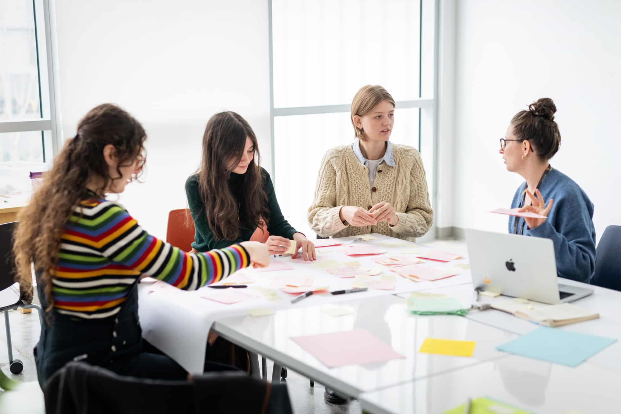 Four students sit around a bright table covered with sticky notes, laptops, and pens, discussing ideas and working collaboratively.