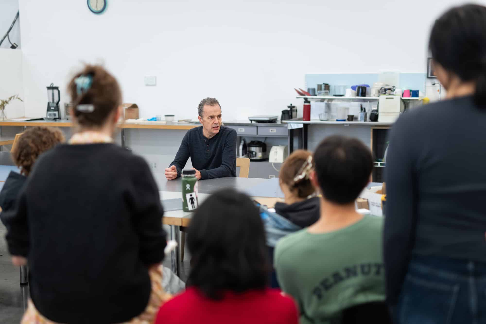 An instructor sits at the front of a studio classroom, speaking to a group of students who are gathered around him.
