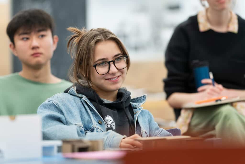A student wearing glasses and a denim jacket smiles while listening in class, with two classmates visible in the background.