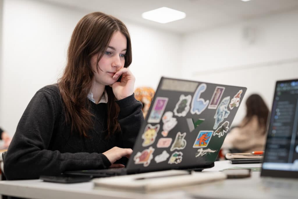 A student sits at a desk, focused on their sticker-covered laptop in a classroom environment.