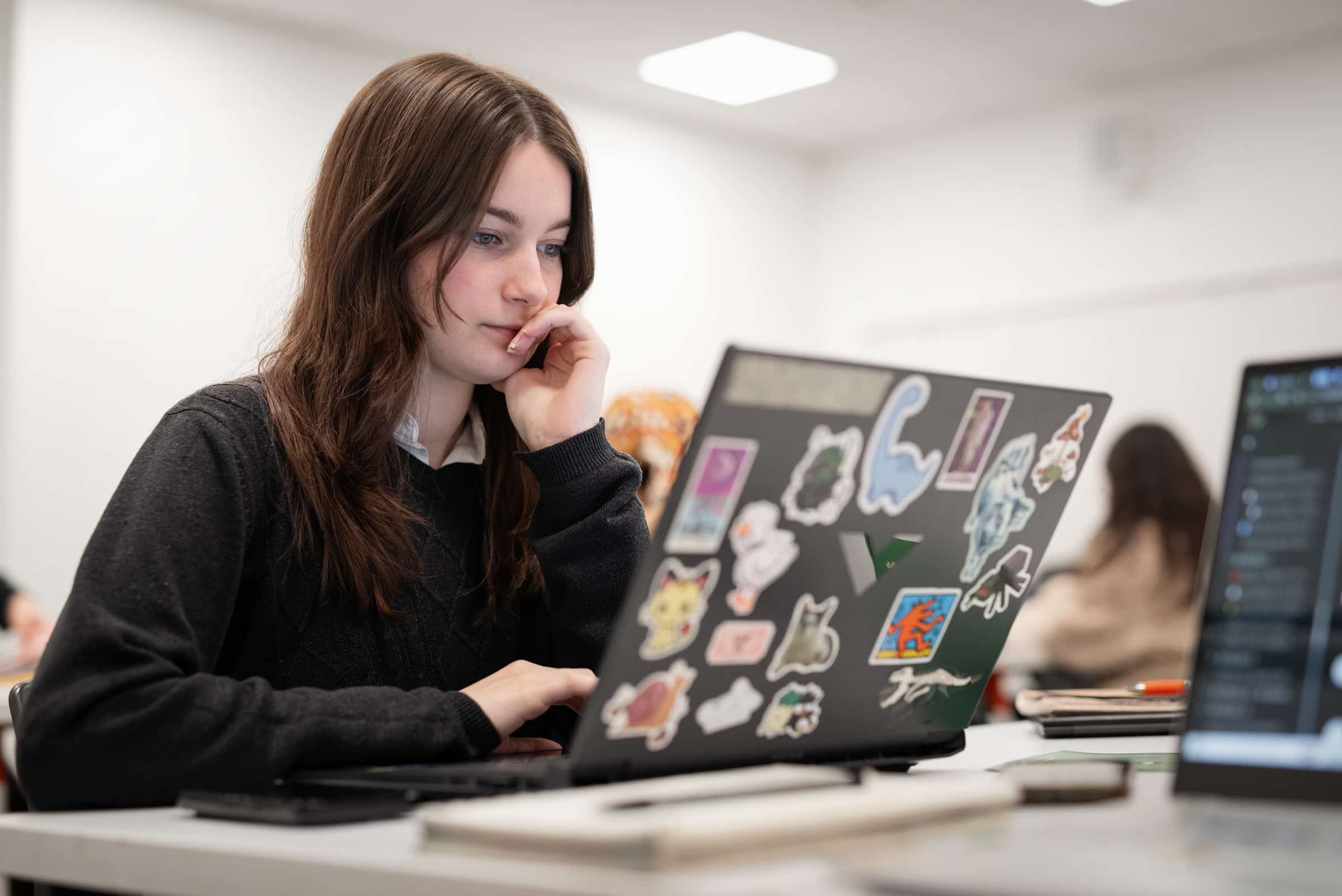 A student sits at a desk, focused on their sticker-covered laptop in a classroom environment.