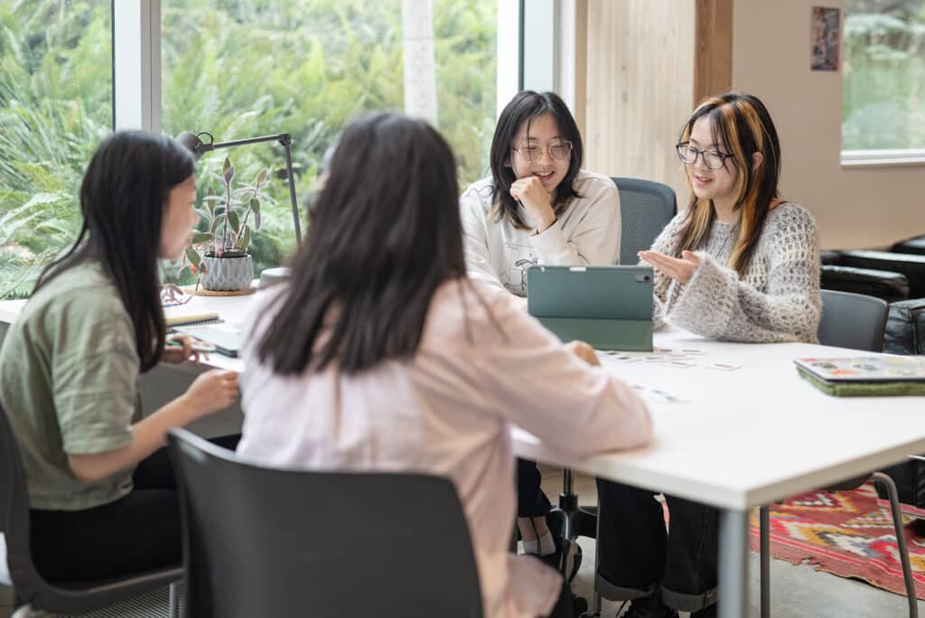 Four students sit together at a table, talking and working on an iPad in a bright lounge with large windows.