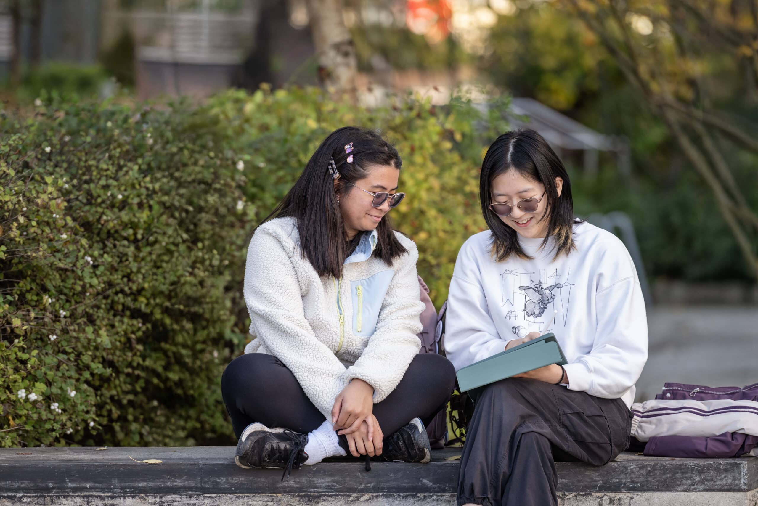 Two students sit outside on campus, smiling and looking at a tablet together.