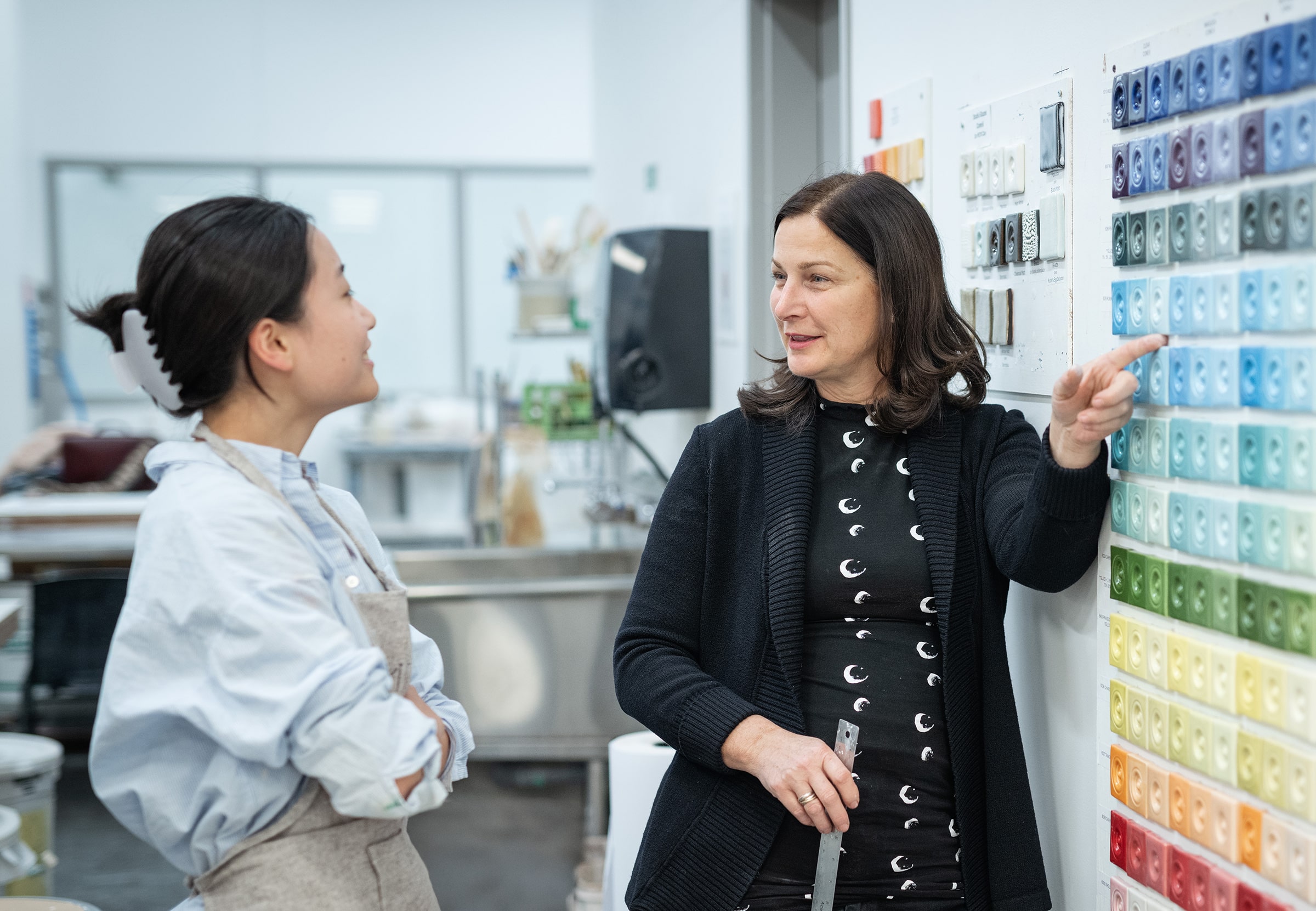 An instructor and a student talk in a ceramics studio beside a wall display of colorful glaze tiles arranged in a gradient. The instructor points to a section of blue tiles while the student listens and smiles, wearing a light blue shirt and apron.