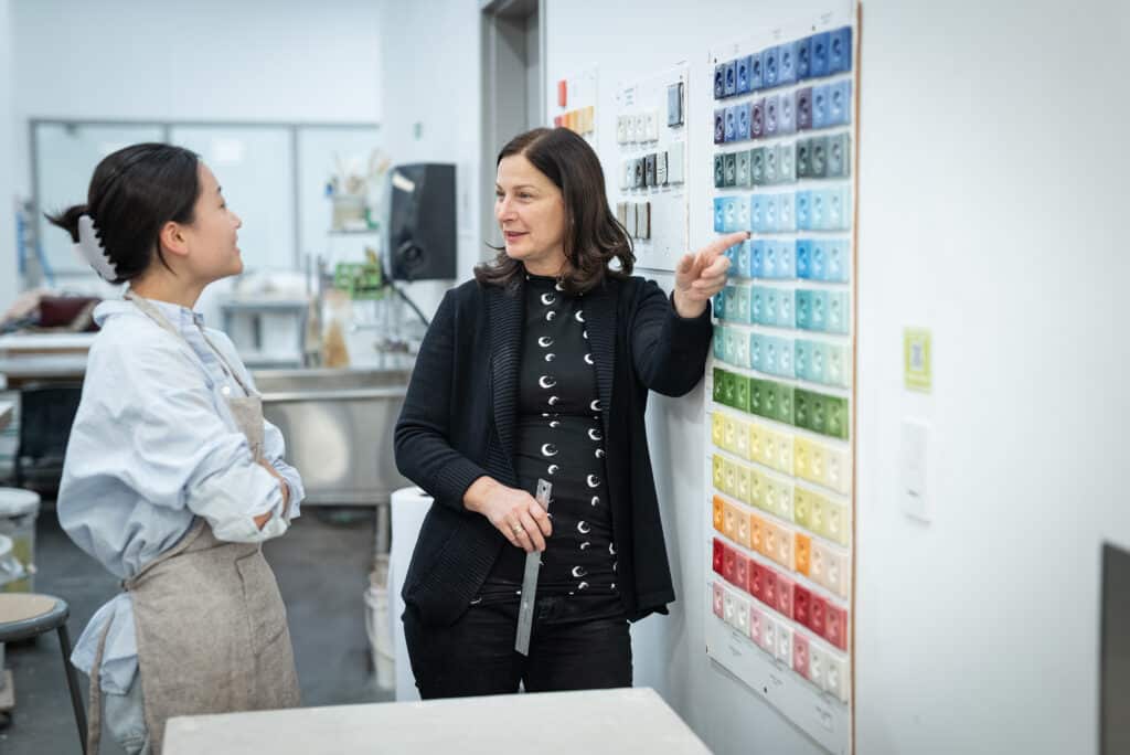 An instructor points to a wall of colourful ceramic glaze tiles while talking with a student in a studio setting.
