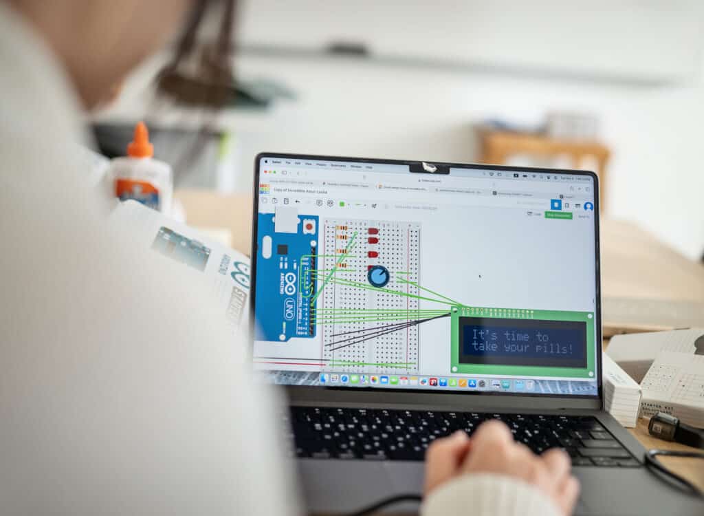 Laptop with Arduino simulation — Close-up of a student working on a laptop screen displaying an Arduino breadboard circuit simulation with the text “It’s time to take your pills!” on a digital display.