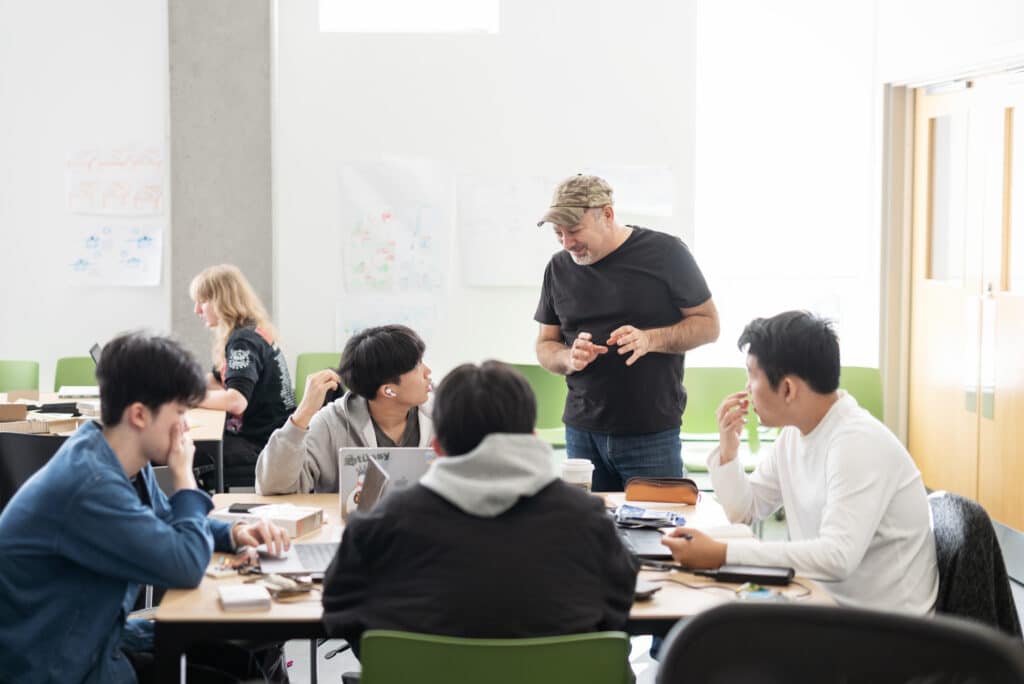 A group of students sit at tables with laptops and notebooks while an instructor in a cap and black shirt gestures as he speaks to them.
