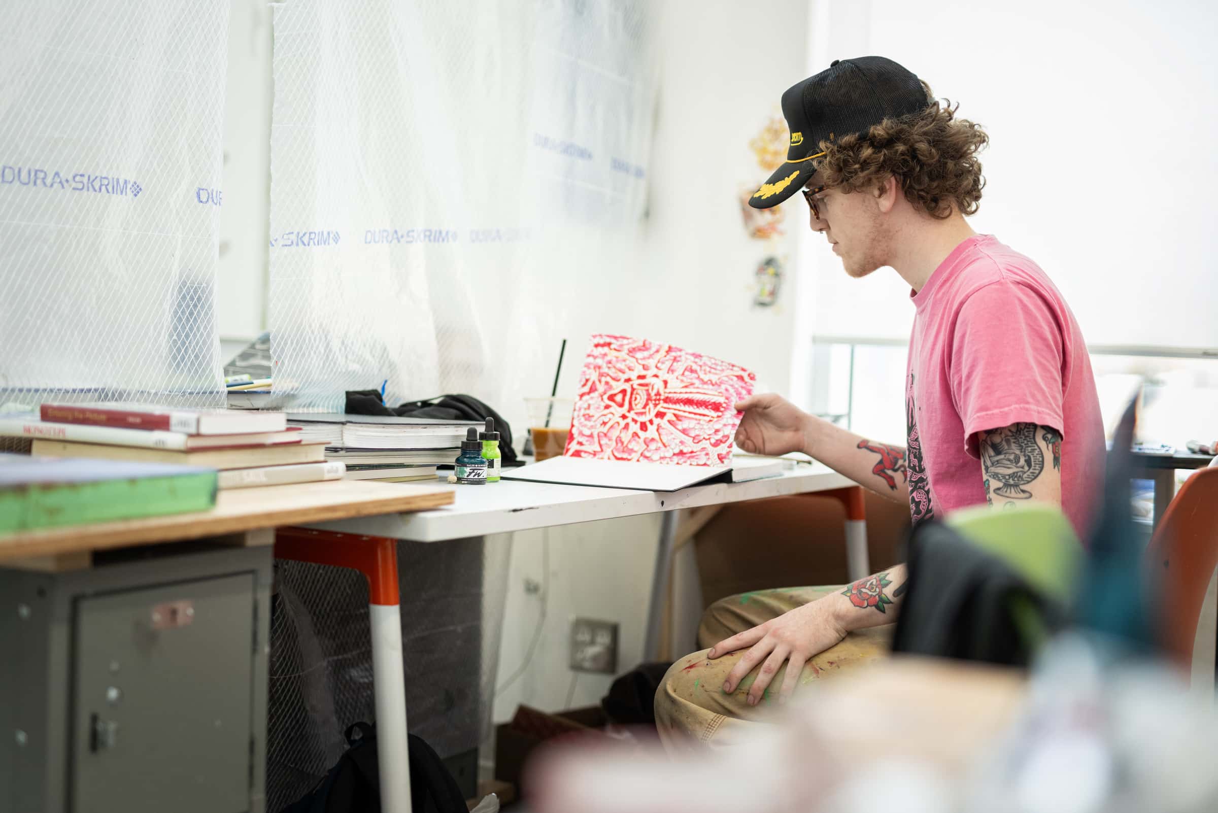 A student in a pink shirt sits at a studio desk examining a vibrant red artwork, surrounded by books and art supplies.
