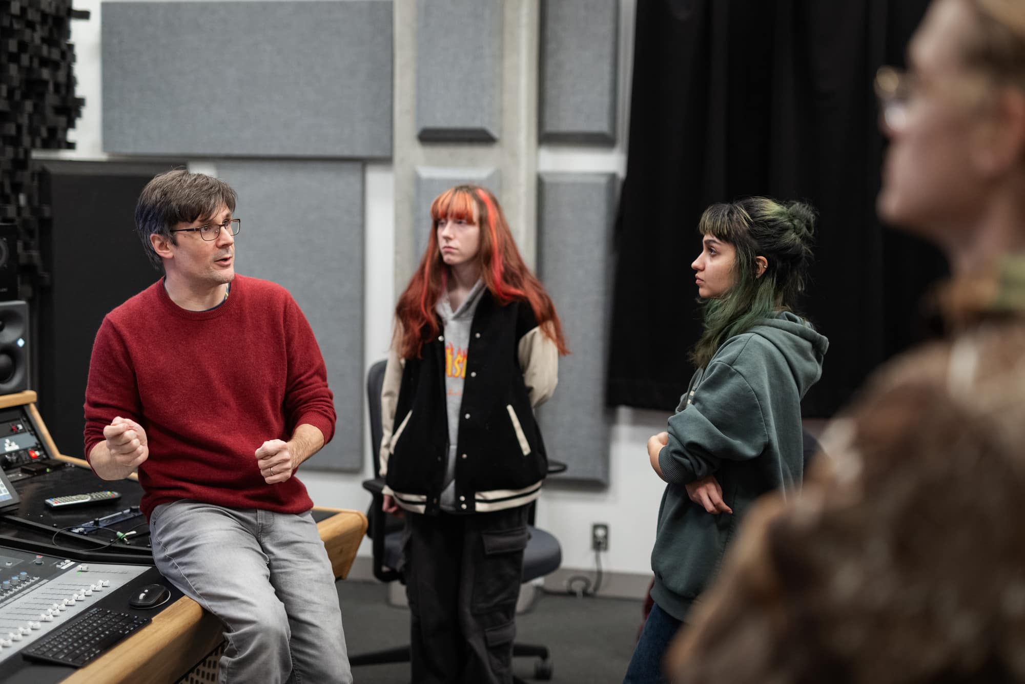 An instructor in a red sweater points toward a screen in a sound studio while students listen attentively.