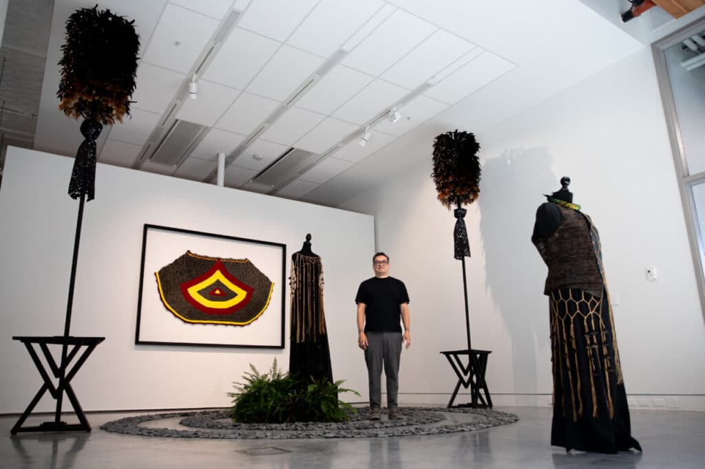 An artist stands in a gallery installation featuring garments, feathered sculptures, and a large wall hanging in red, yellow, and black.