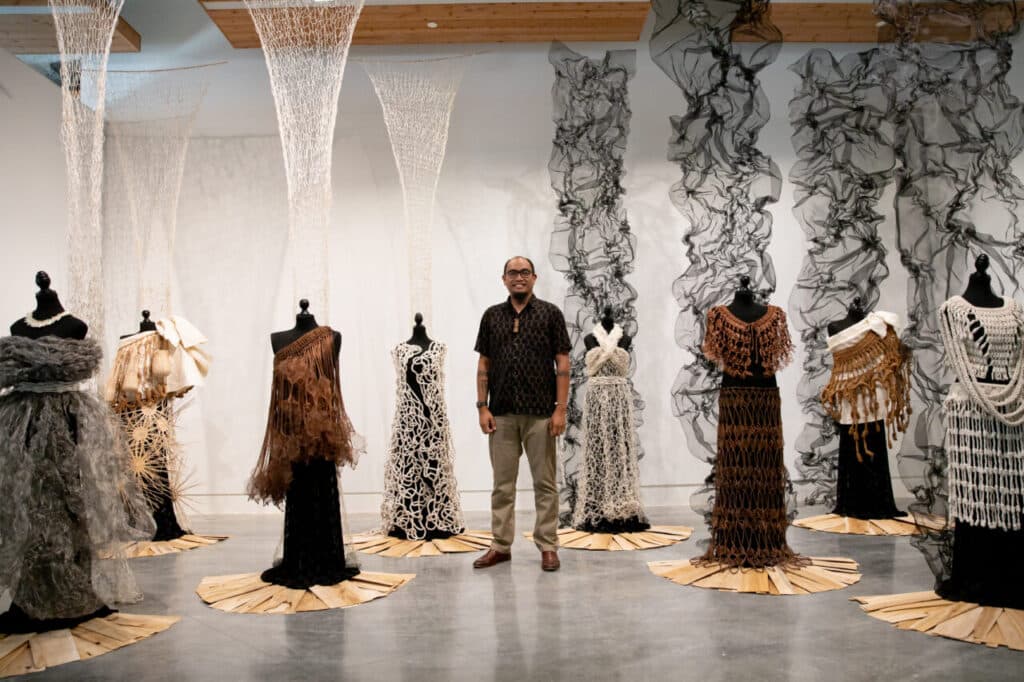 An artist stands among a group of mannequins dressed in intricately woven and knotted garments made of natural fibres, displayed in a gallery with sheer hanging textiles.