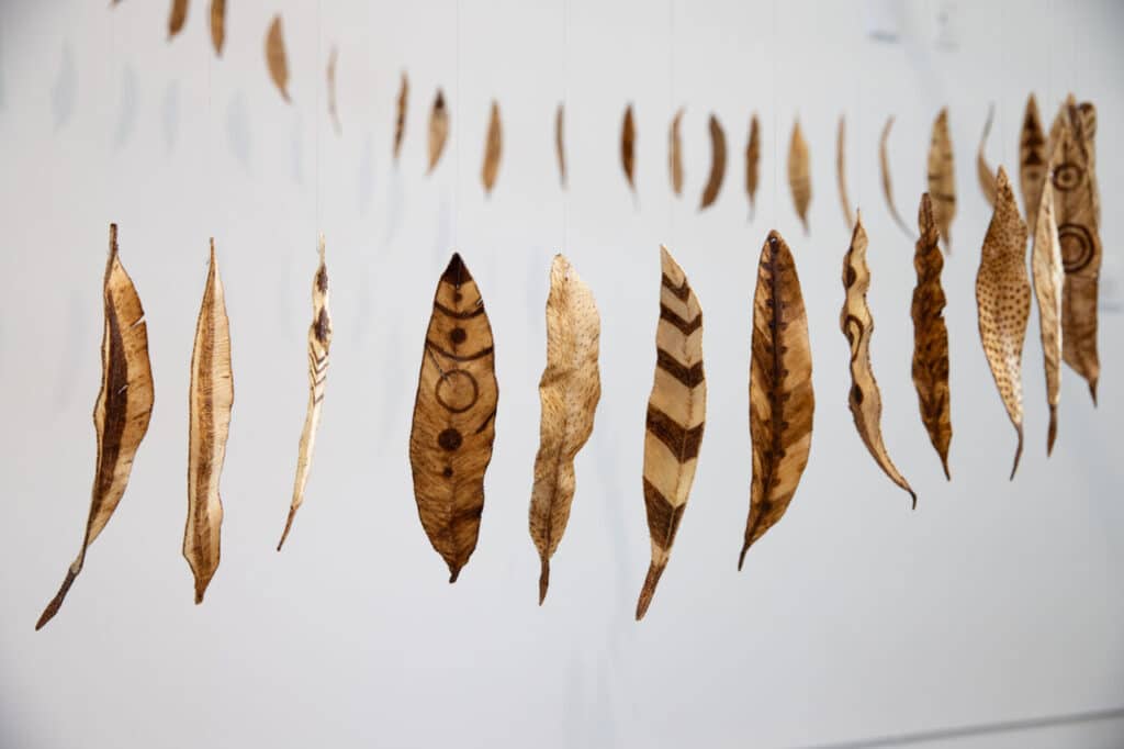 A hanging installation of feather-shaped forms made from natural materials, each etched or burned with unique geometric and organic patterns.