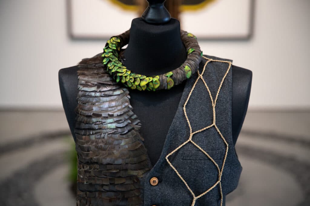 A close-up of a black mannequin wearing a dark textured vest adorned with layered brown scales, green accents, and netted gold rope.