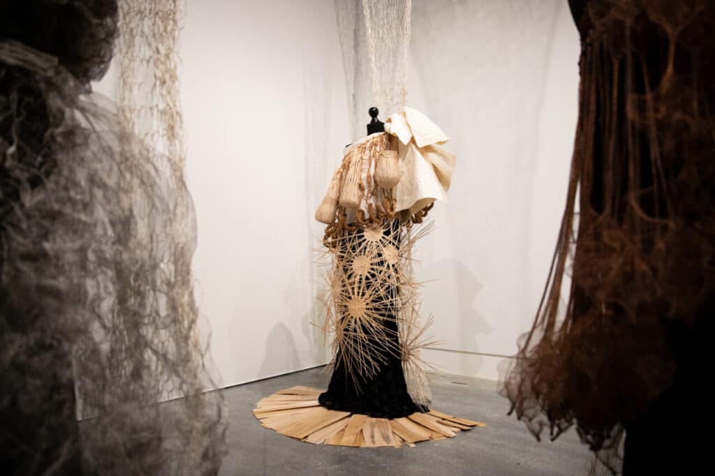 An installation view of a woven garment on a mannequin with layered textures of natural fibres and paper-like materials, surrounded by hanging netted sculptures.