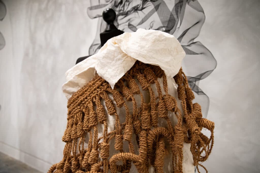A close-up of a sculptural garment on a black mannequin, featuring a woven brown rope structure layered over textured white fabric.