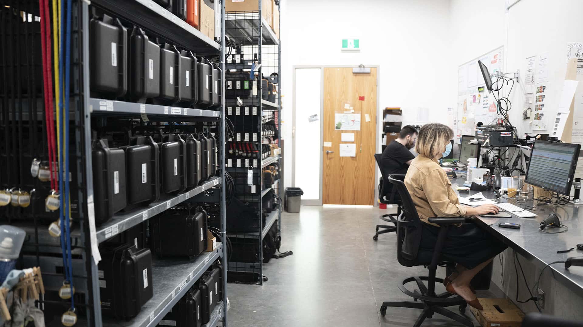 A storage room with rows of black equipment cases on shelves, and two people working at computer desks along the wall.