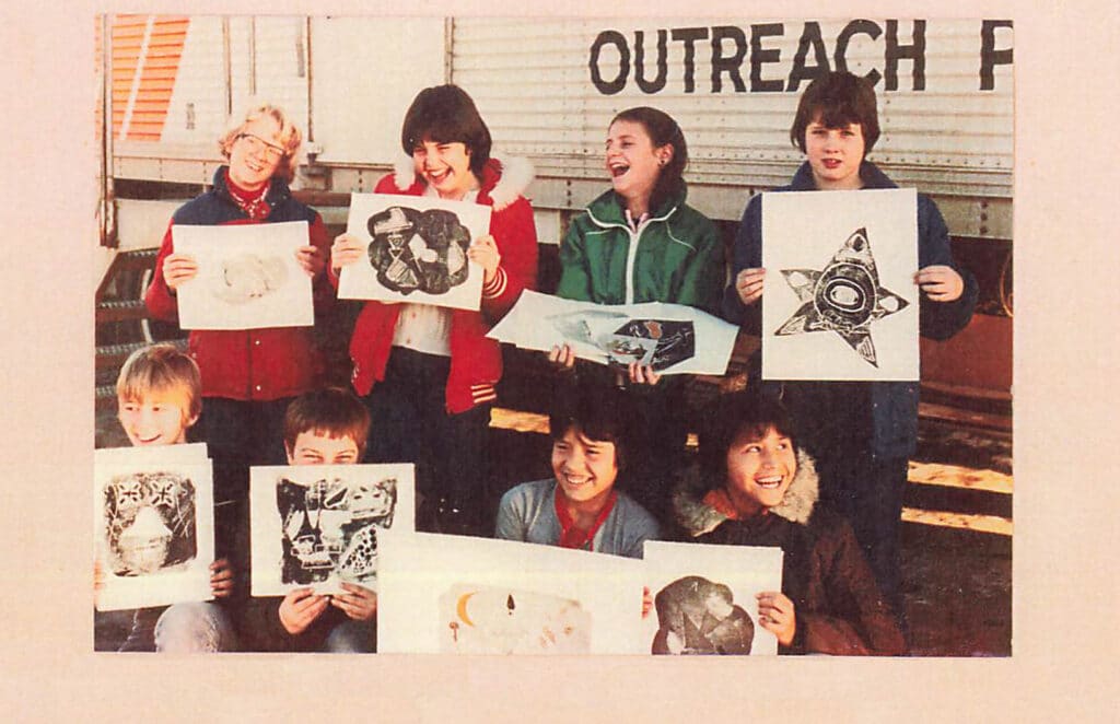 A group of smiling children hold up their artwork while standing outside a mobile art studio.