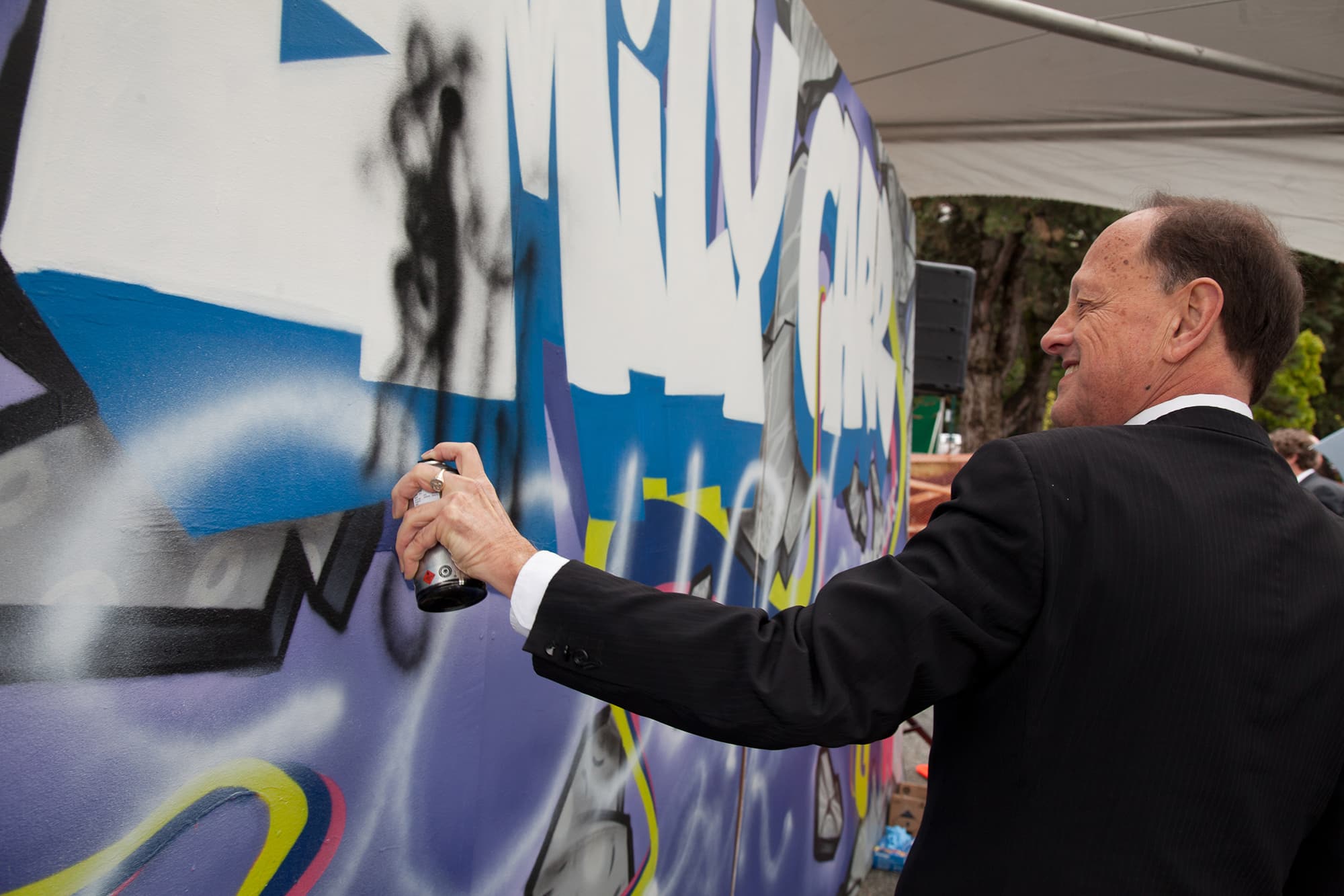 A man in a suit spray paints a large colorful mural featuring graffiti-style text that reads “Emily Carr.” The mural is outdoors under a white tent, and the man smiles as he adds paint to the artwork.