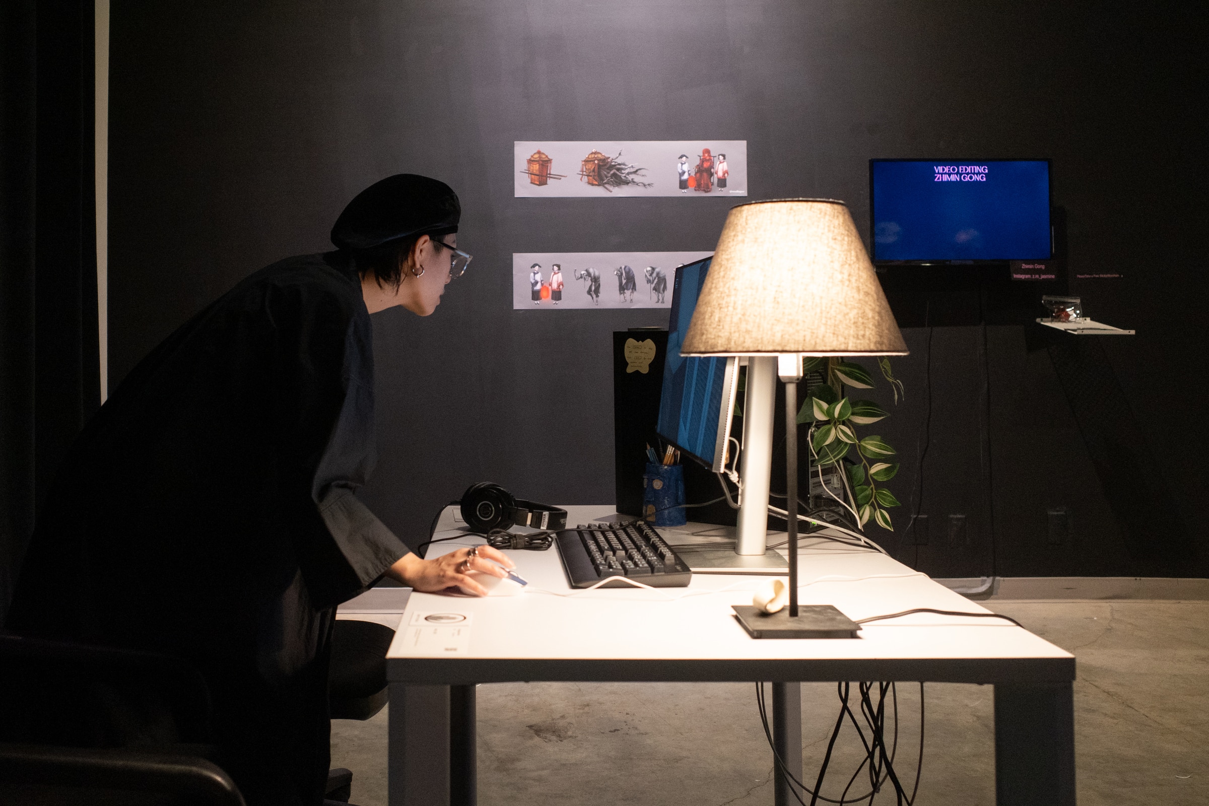 A person leans over a desk illuminated by a lamp while viewing digital artwork in a dark exhibition space.