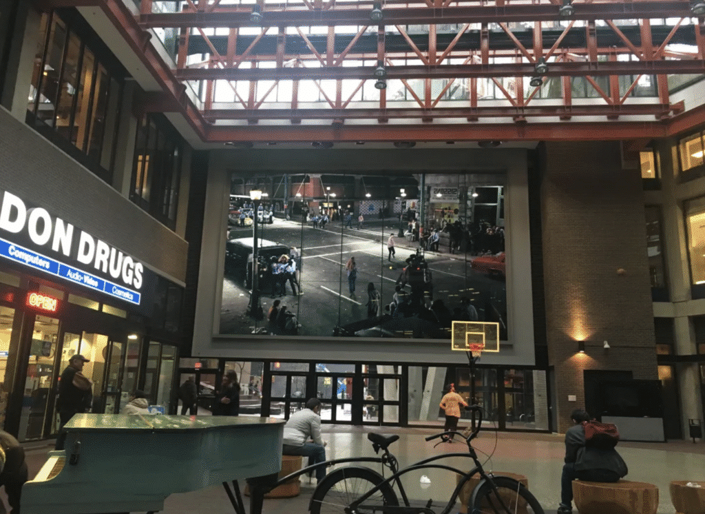 A large-scale artwork is displayed inside an indoor atrium where people are gathered near shops and seating.