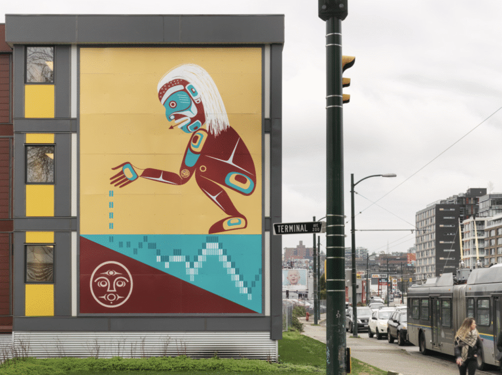 A large mural on a building depicts an Indigenous figure extending a hand, designed in a Northwest Coast style.