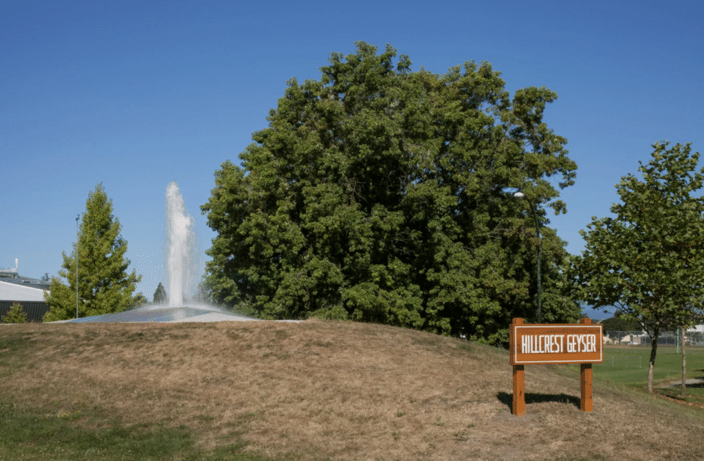 A water fountain shoots a tall plume into the air from a grassy mound beside a sign reading “Hillcrest Geyser.”