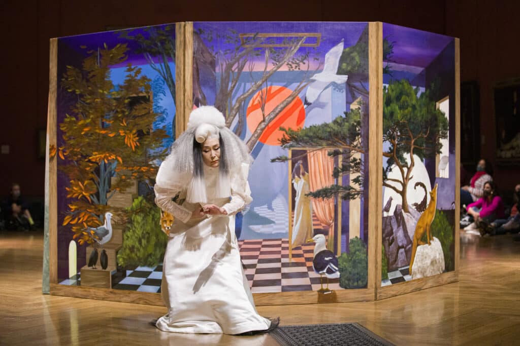 A performer dressed in white kneels in front of a painted backdrop featuring vivid landscapes and animals.