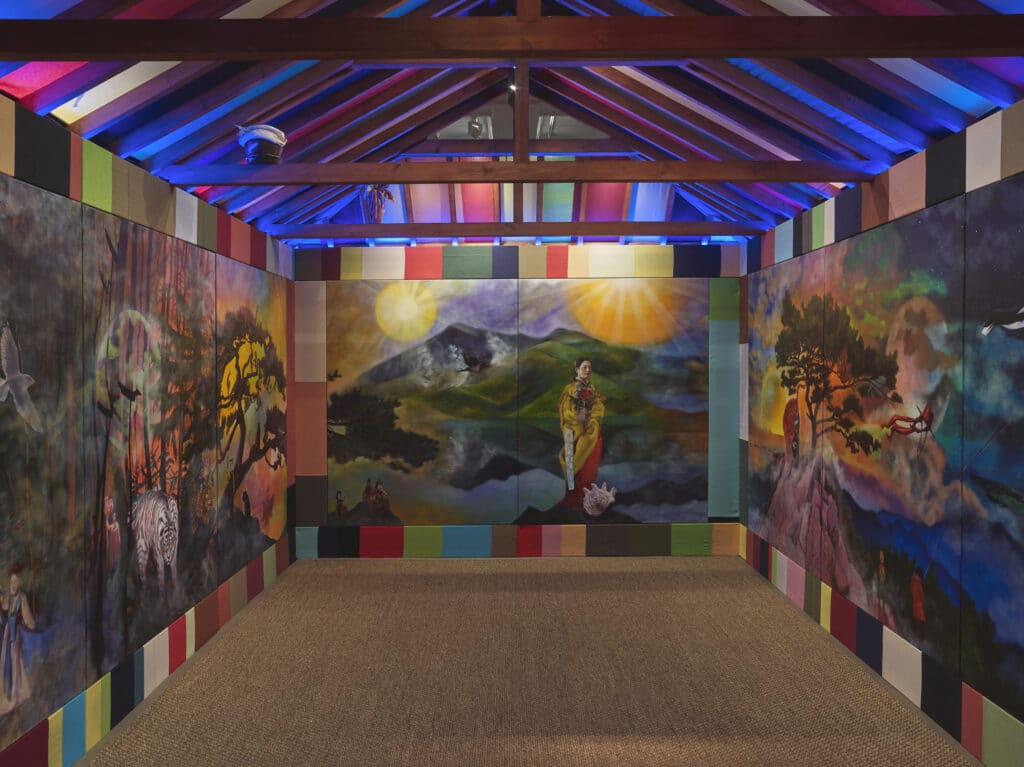 A colourful gallery installation features large painted panels depicting surreal landscapes, animals and human figures under vibrant lighting.