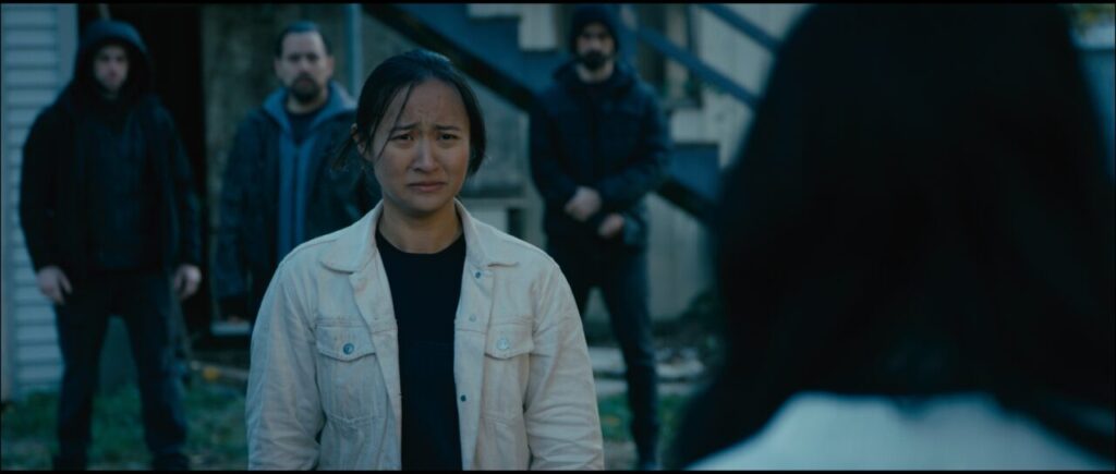 A person stands outside facing someone off-screen, with three people in dark clothing watching from behind.