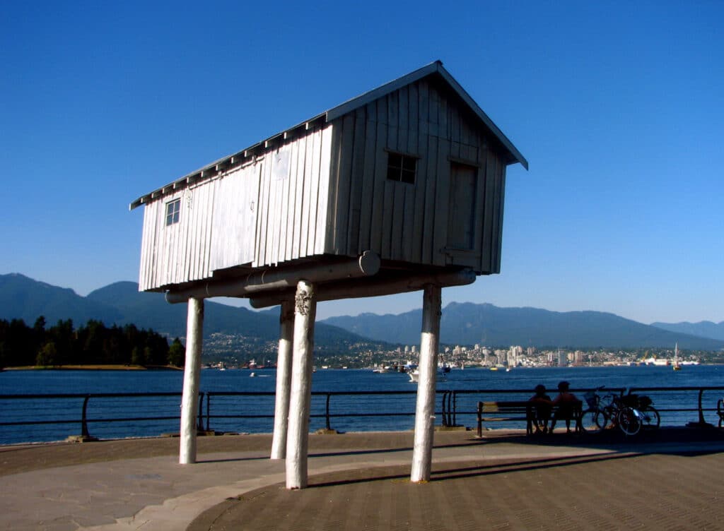 A small silver-grey shed-like sculpture on stilts stands above the seawall with mountains and city skyline behind it.