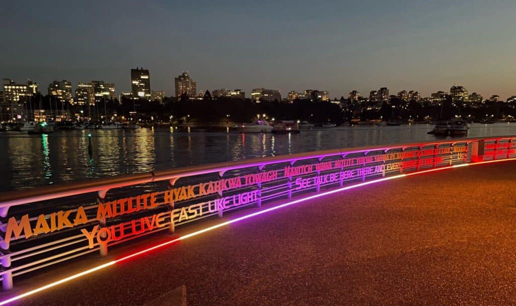 Illuminated text in multiple colours runs along a waterfront railing at night, reflecting on the water.
