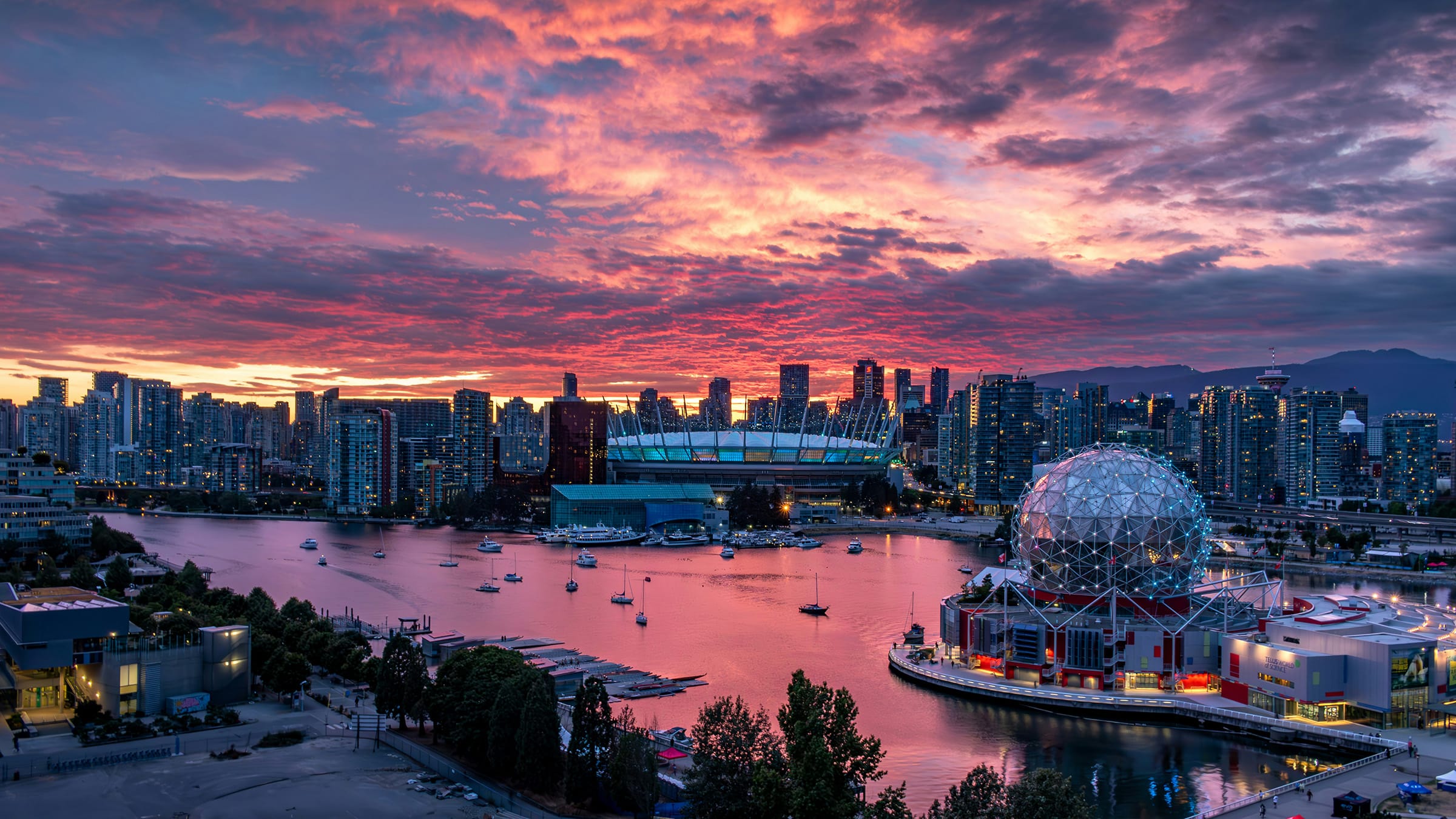 A panoramic view of downtown Vancouver at sunset, with BC Place Stadium and Science World glowing beside False Creek under a dramatic pink and purple sky.