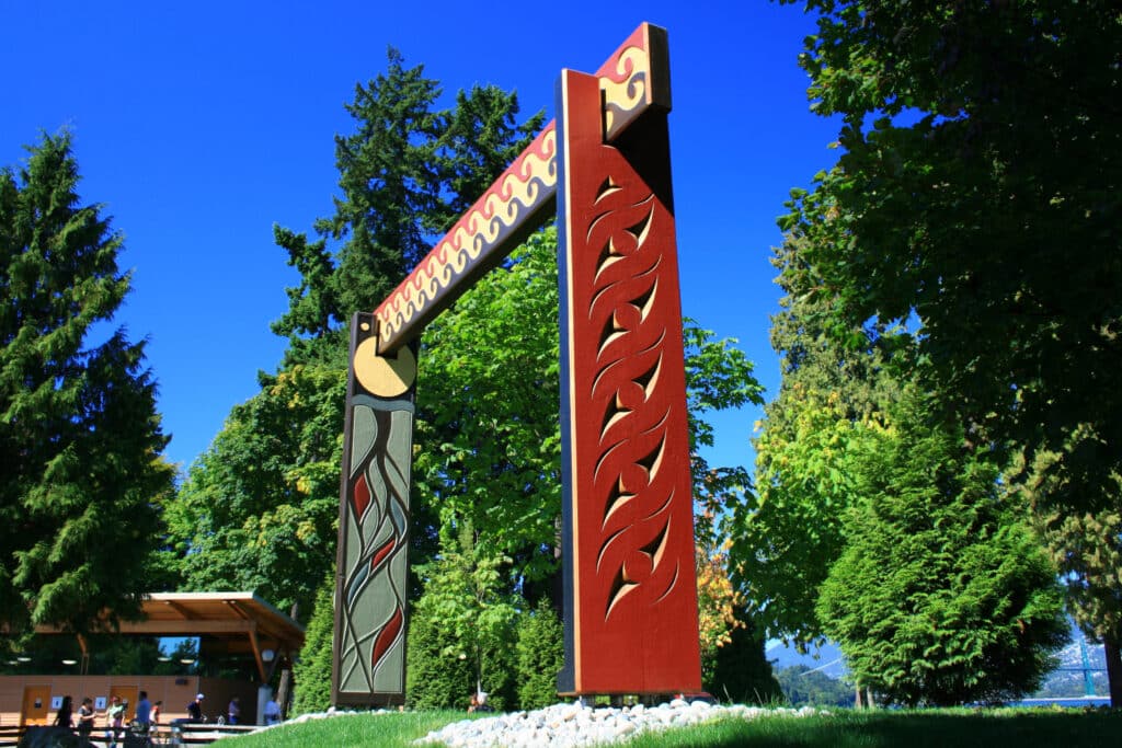 A large red and green carved gateway with Coast Salish designs stands among tall trees in a park.