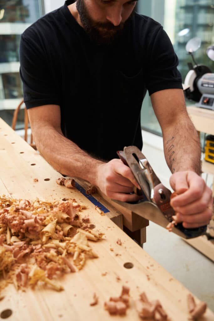 A person in a black shirt using a hand plane to smooth a piece of wood at a workbench, with piles of wood shavings scattered across the surface.