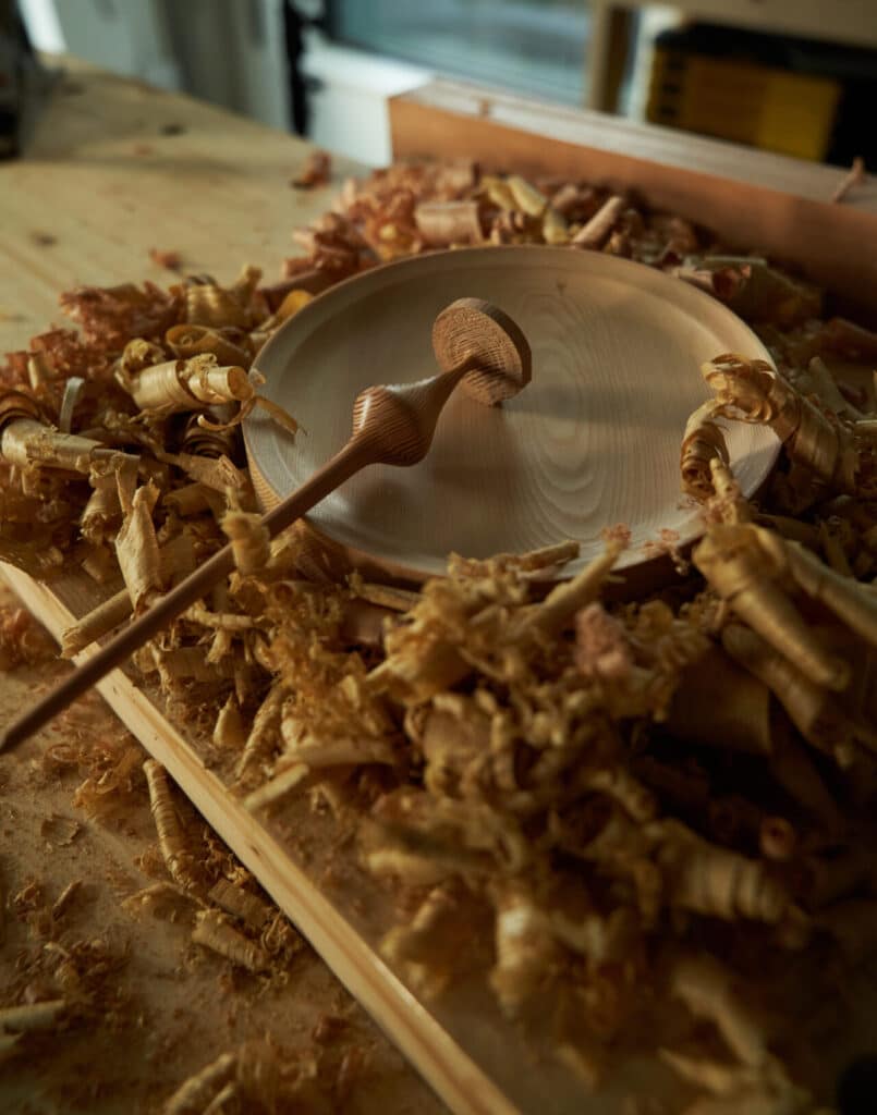 A round wooden plate surrounded by curled wood shavings on a workbench, with a wooden turning tool resting across the plate.