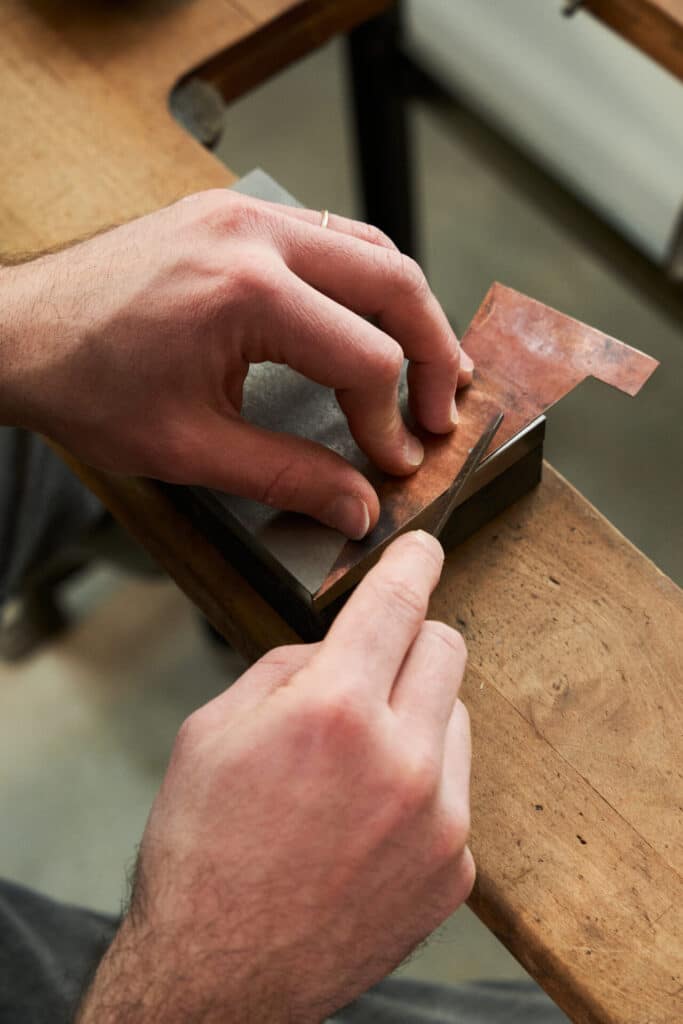 Close-up of a person’s hands sharpening a small tool on a whetstone, with a piece of copper held steady for precision work.