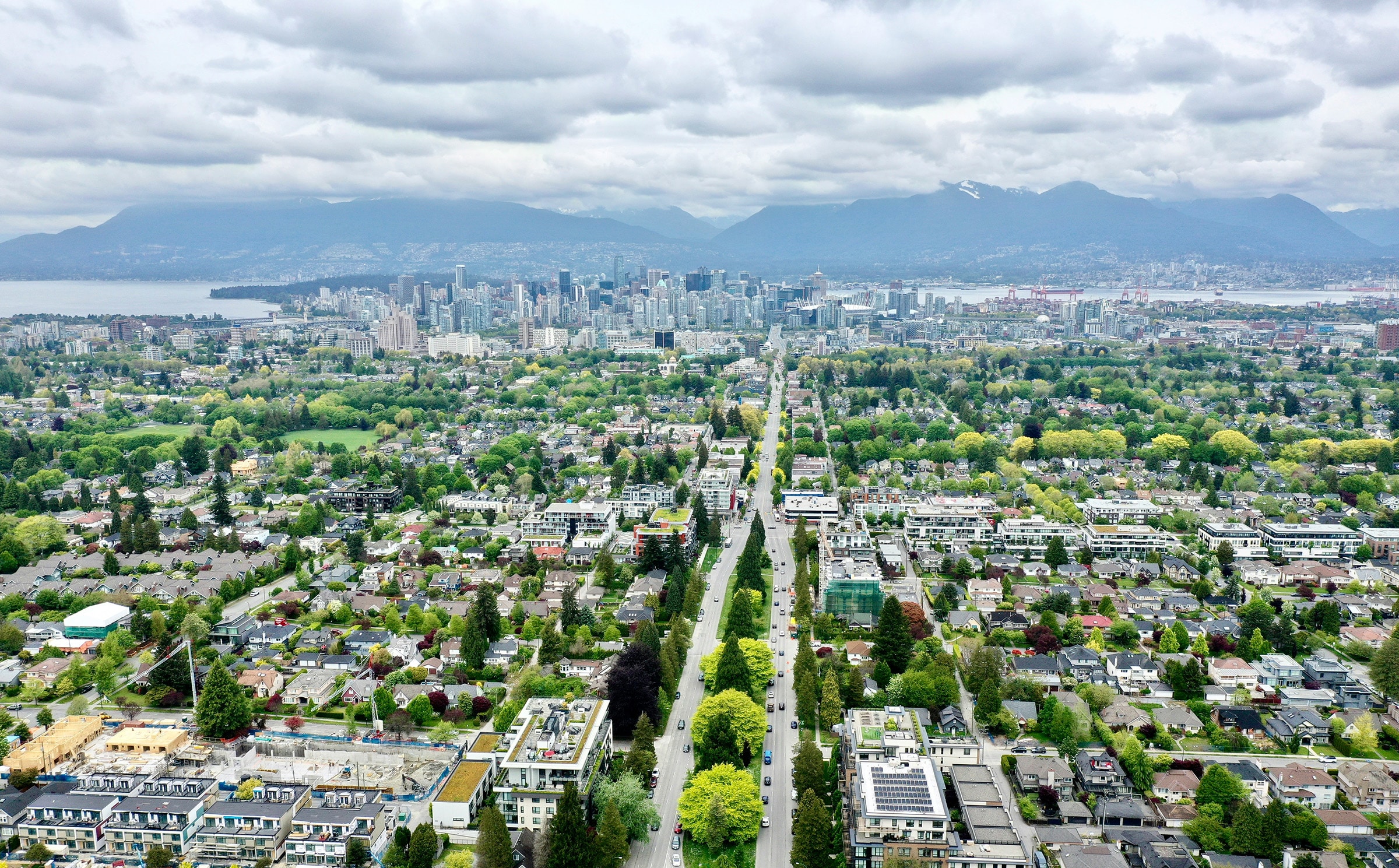Aerial view of Vancouver’s tree-lined streets and residential neighborhoods leading toward the downtown skyline and mountains under a cloudy sky.