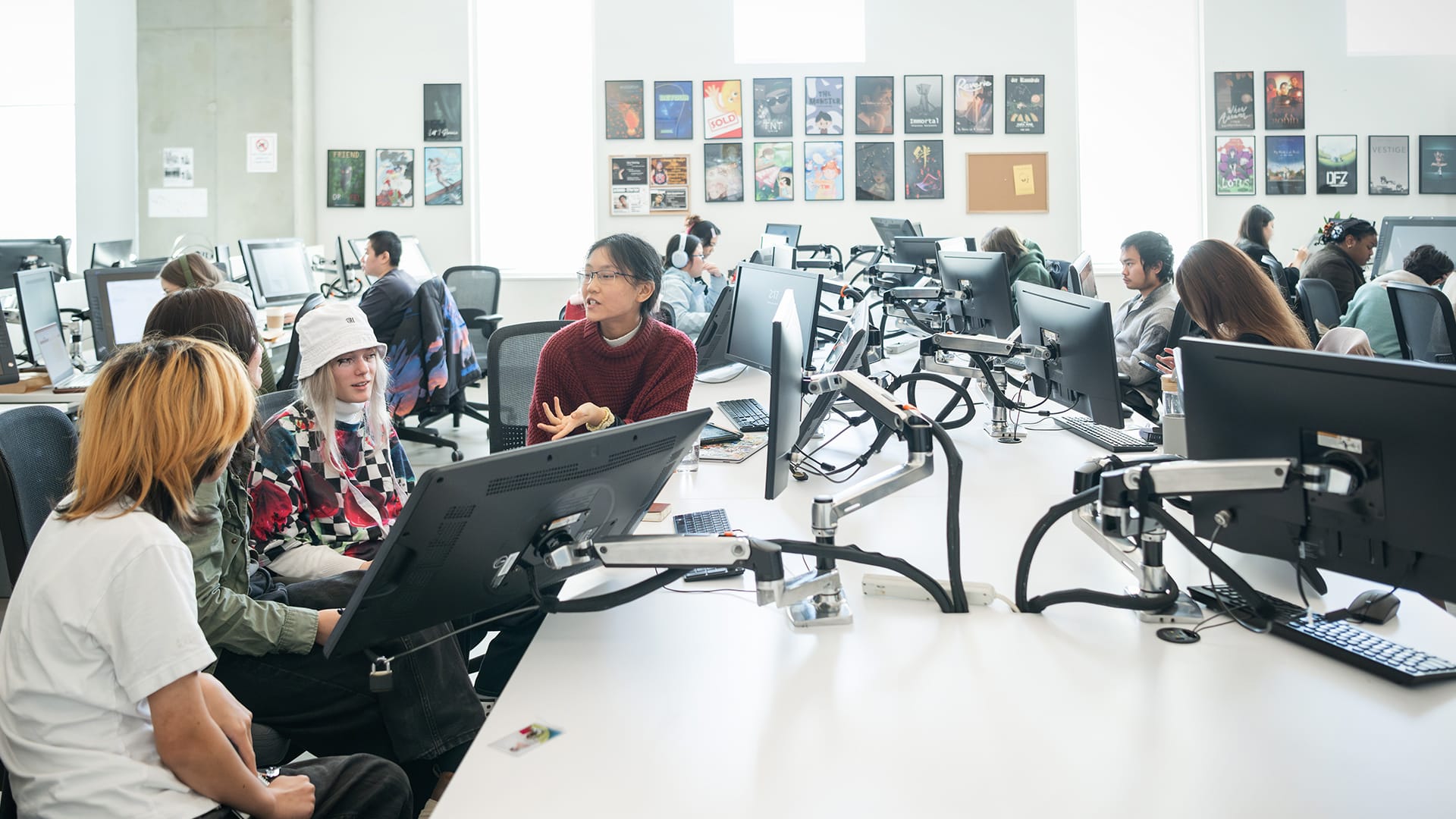 A group of students collaborates around a workstation in a bright, modern computer lab. One student in a red sweater gestures while speaking to peers — including one with platinum hair wearing a white bucket hat and another in a white t-shirt — as they all focus on a shared monitor. Rows of computers line the room, with other students working independently in the background. The walls are decorated with colorful posters and artwork, reinforcing the creative atmosphere of the space.