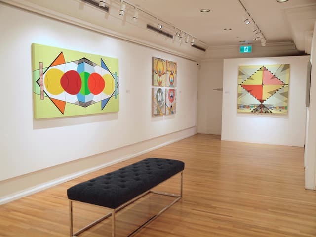 Bright gallery interior featuring geometric abstract paintings in vivid colours on white walls, with a dark bench centred on a polished wood floor.