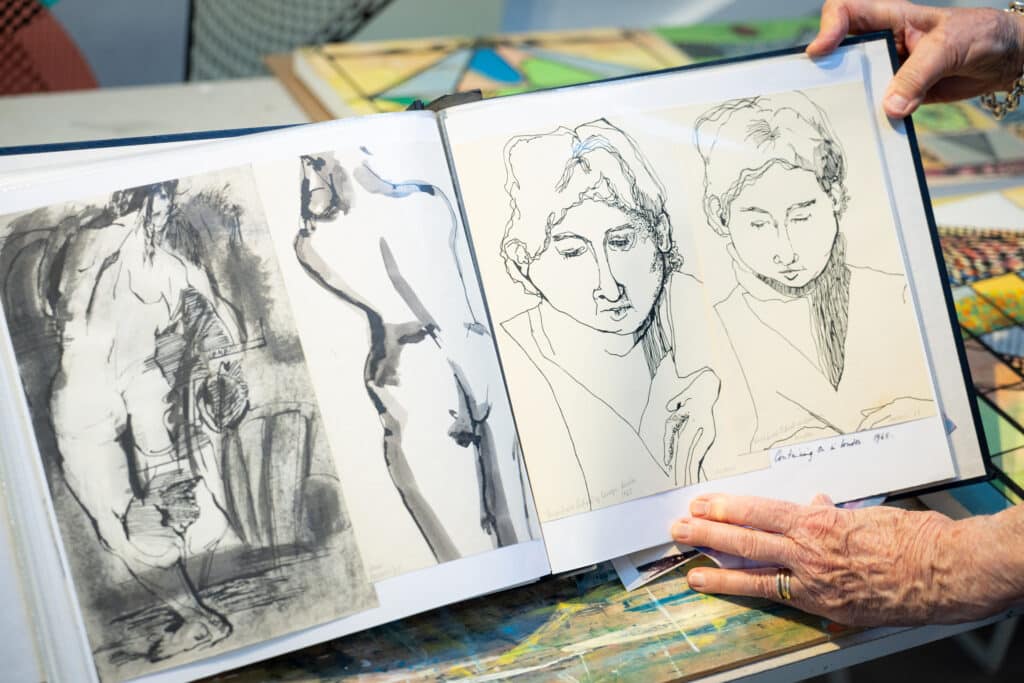 Close-up of hands holding a sketchbook open to figure and portrait drawings in ink and charcoal on a studio table.
