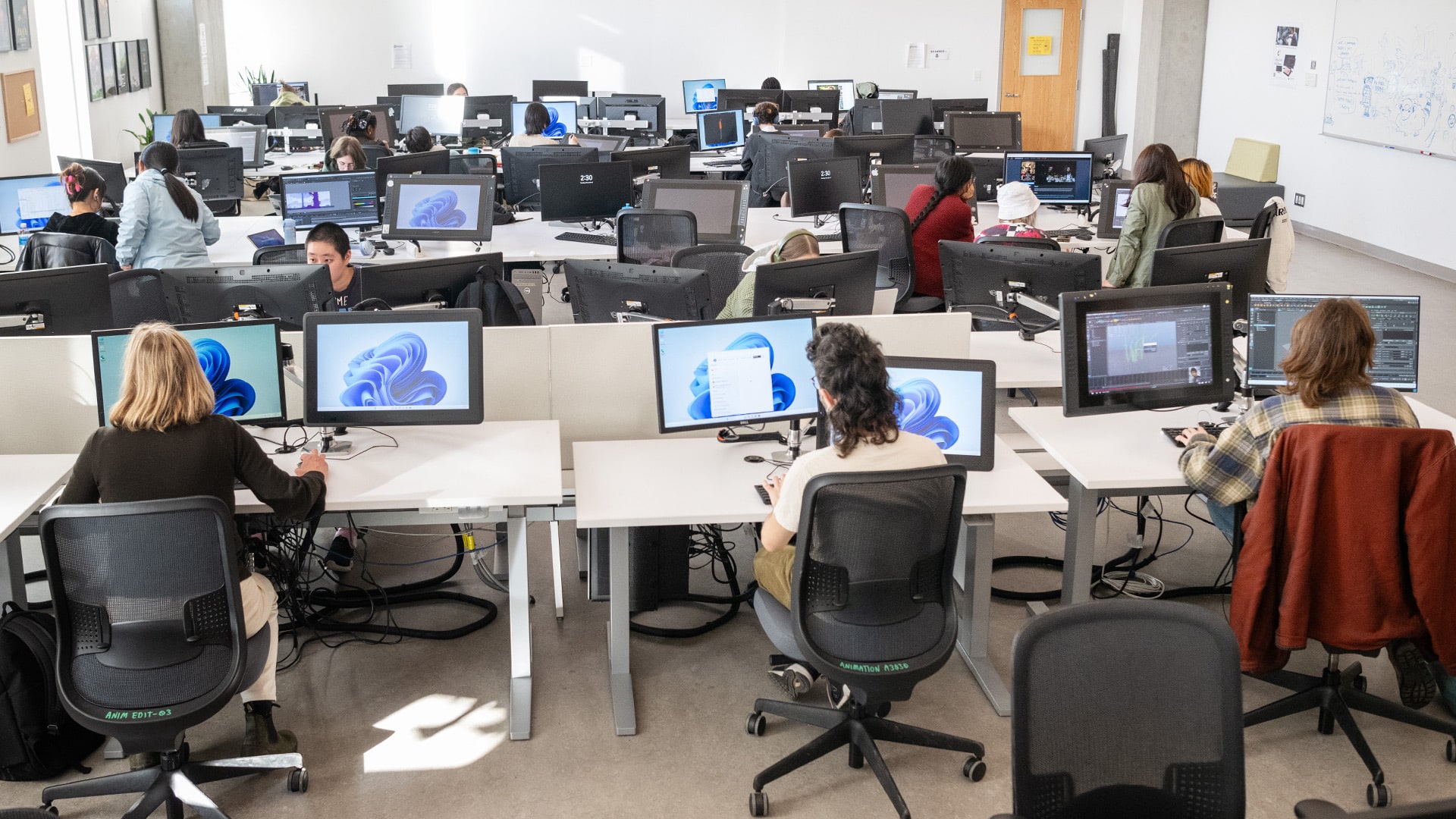 A wide-angle view of a bright, modern computer lab at Emily Carr University of Art + Design, where students are seated at individual workstations learning 3D software. Most screens display some showing its blue swirl splash screen, others with 3D modeling or animation tools active. Students are focused on their monitors, some wearing headphones, surrounded by black mesh chairs and white desks. Sunlight streams in from large windows, illuminating the collaborative, tech-driven learning environment.