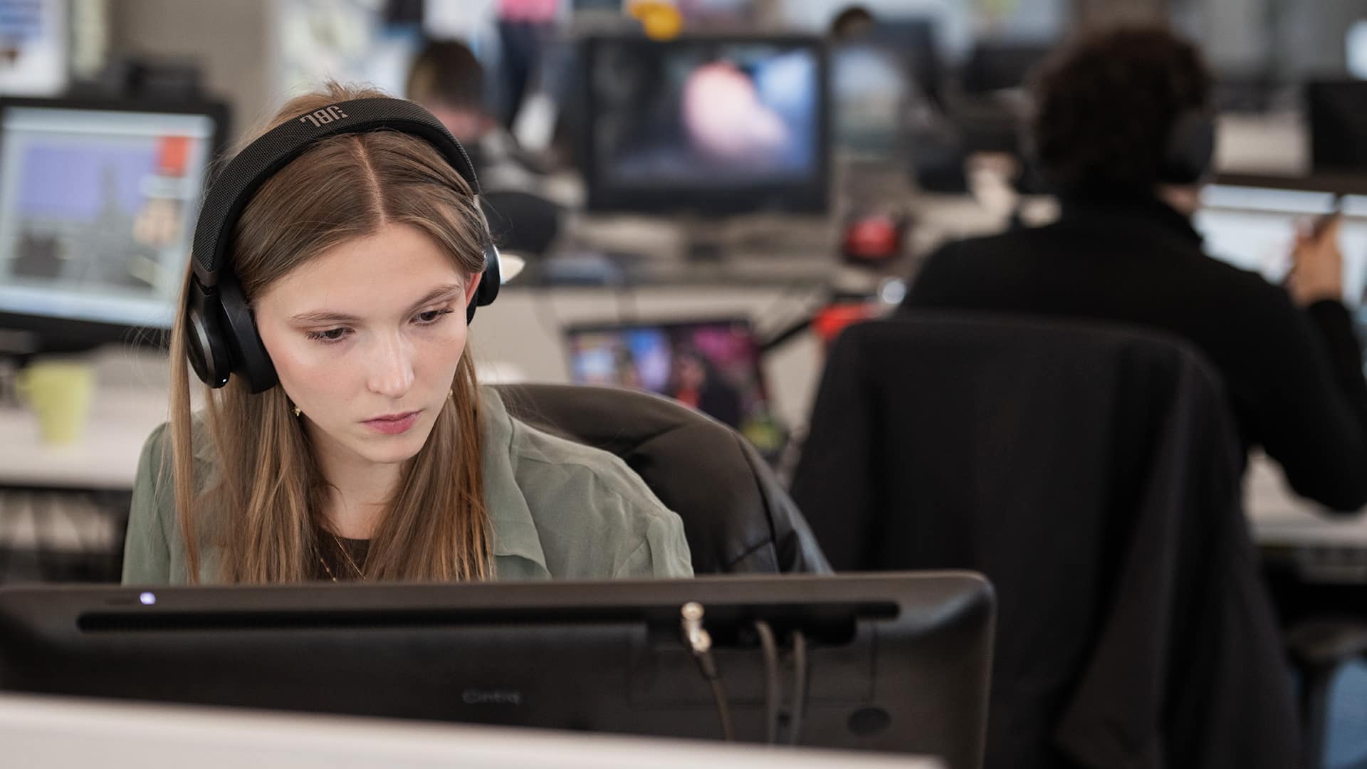 A focused student wearing black JBL over-ear headphones works intently at a computer in a busy studio or classroom. She has long brown hair and wears a light green shirt, her gaze fixed on the screen. In the blurred background, other students are seated at desks with monitors, suggesting a collaborative or instructional environment.