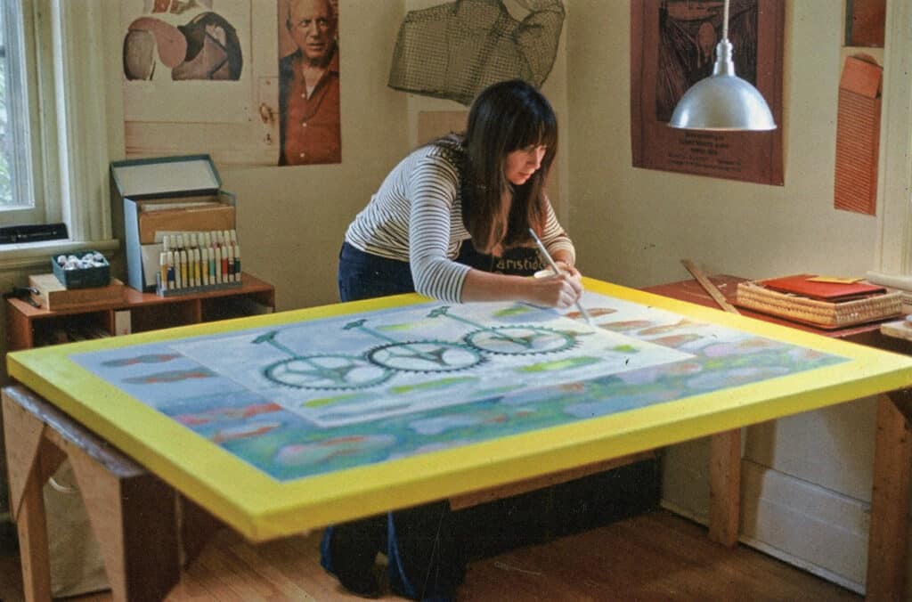 Artist painting a large colourful canvas on a table in a studio filled with posters and art supplies.