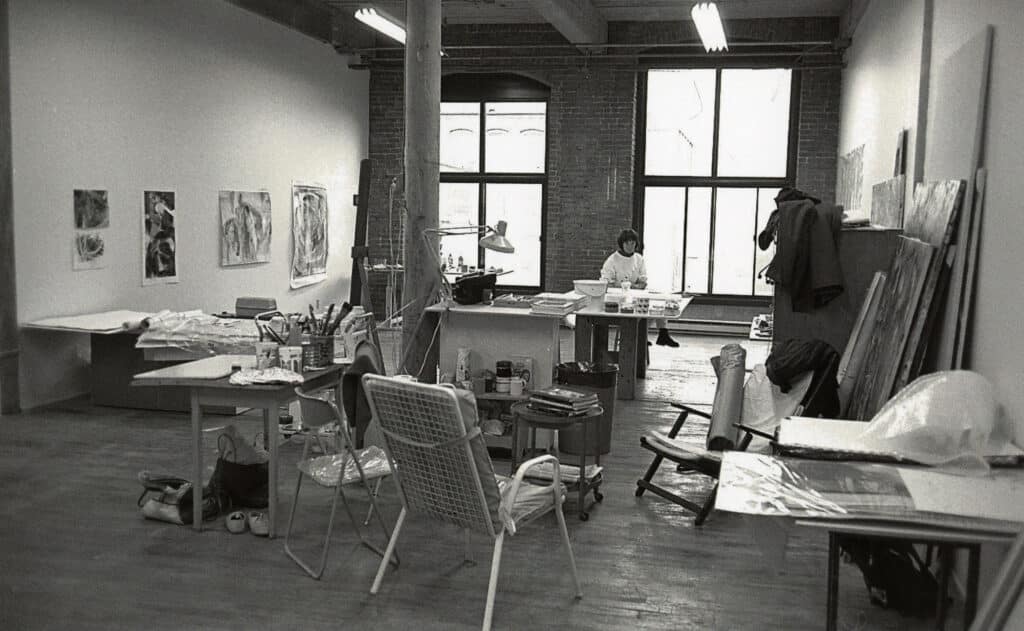 Black-and-white photo of an artist working in a spacious studio with brick walls and large windows.