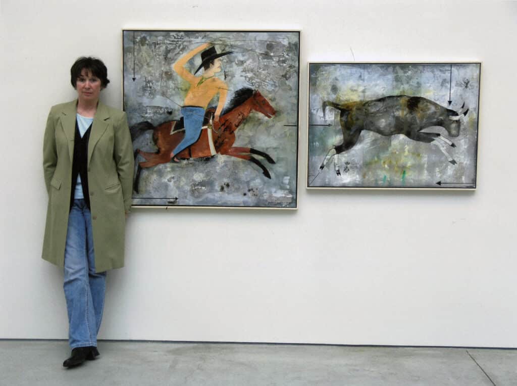 Artist standing beside two paintings depicting a rider on horseback and a running bull.