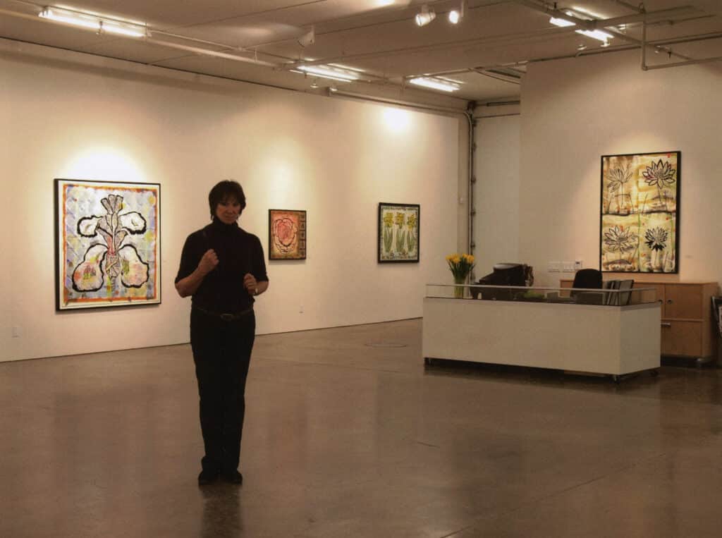 Artist standing in a gallery surrounded by floral paintings and warm lighting.