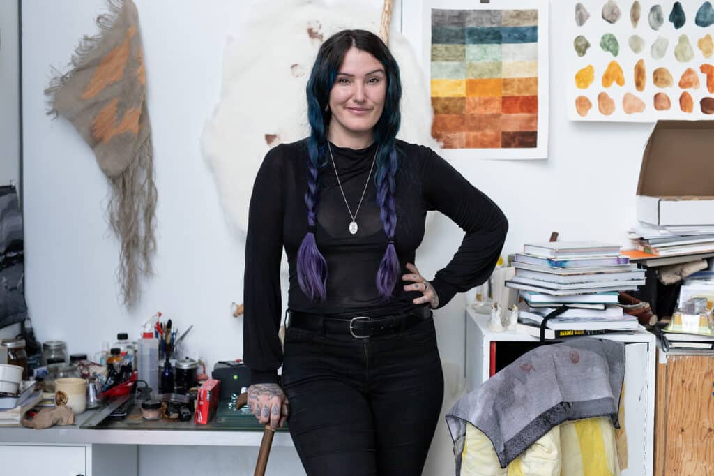 A person with long braids stands in a studio surrounded by art materials, books, and samples of textured and coloured works pinned on the wall.