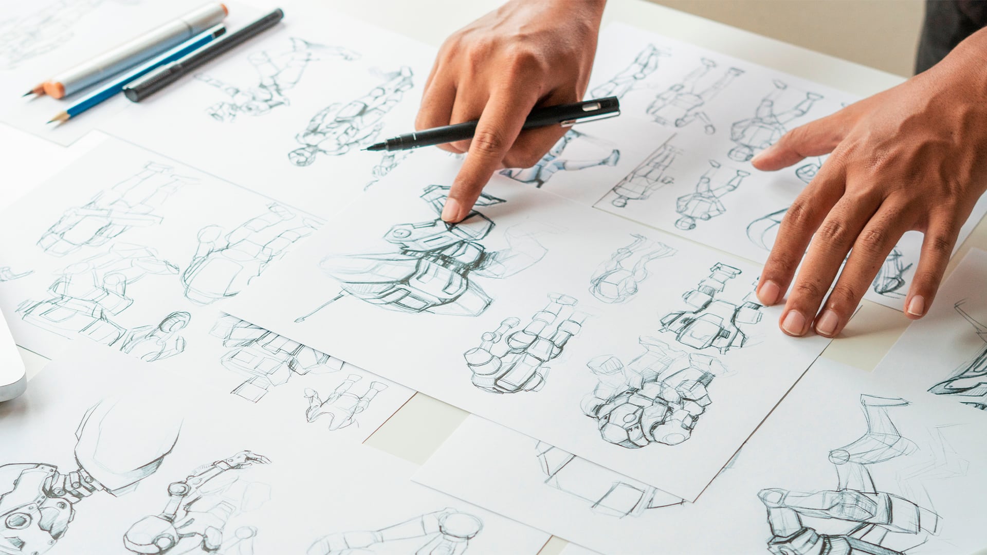 A close-up view of hands working on multiple sheets of paper covered in detailed pencil sketches of robotic or armored figures. One hand holds a black pen, pointing to a specific detail on a central drawing, while the other steadies the page. Several pencils and markers lie nearby on the white desk. The sketches show dynamic poses and mechanical anatomy, suggesting concept development for animation, game design, or character art.
