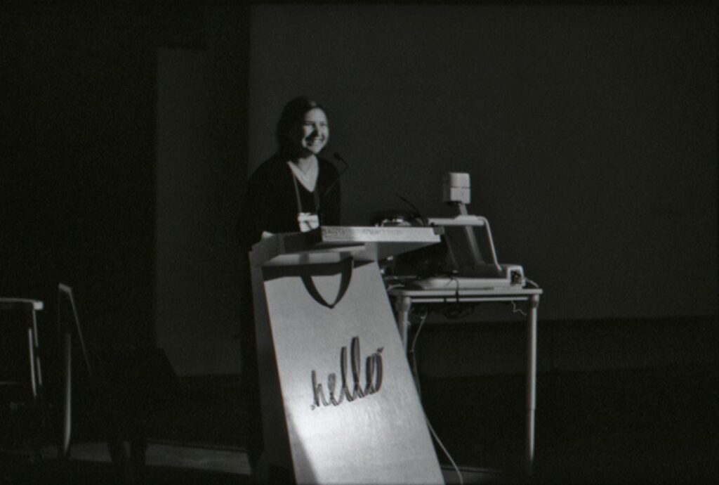 A person stands smiling at a microphone behind a podium against a darkened background. The podium features the word "hello" on its front.